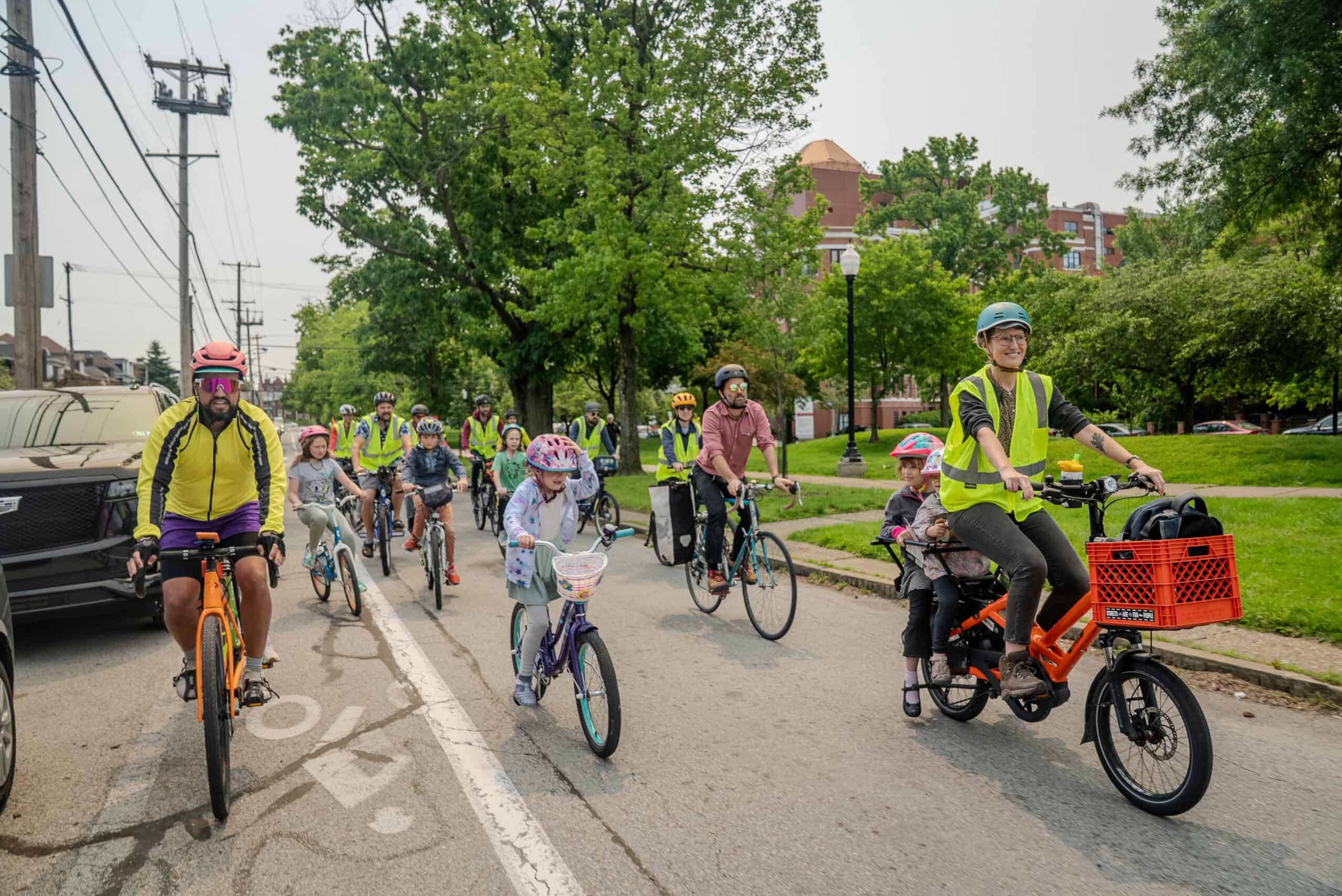 A smiling woman on a cargo bike with a neon vest is riding by a park with her two kids on the back seat. several children and adults are riding around her. The adults have neon safety vests to indicate they are bike bus leaders. 
