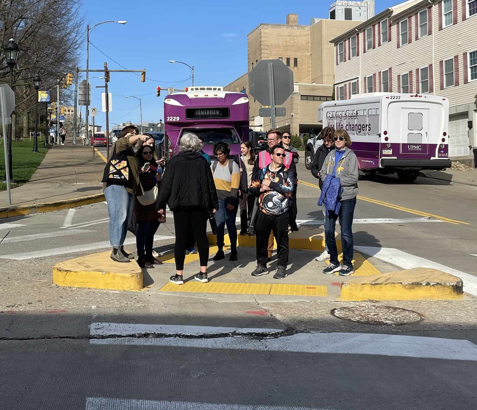A group of 12 people stand at a pedestrian island, while two UPMC shuttles pass nearby.