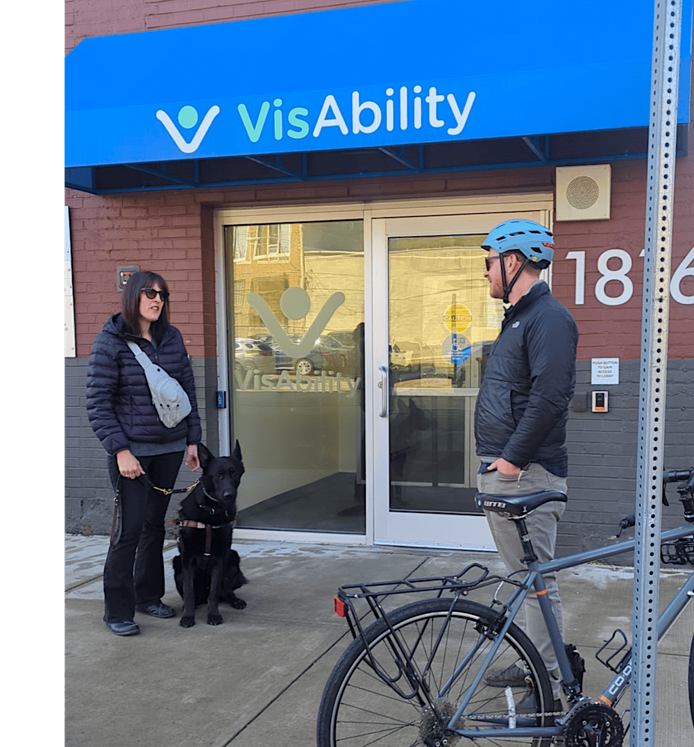 A photo of Seth, a fair skinned man with red hair and wearing a bike helmet, and his bike, standing with Amelie, a fair skinned woman with medium length brown hair, and her black german shepherd guide dog. They are standing outside of VisAbility Pittsburgh and chatting. 