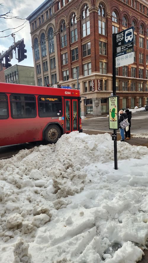 A red bus approaches and people line up to get on, but they are doing so in the street because the entire area around the sidewalk and bus stop is fully covered in mounds of snow.