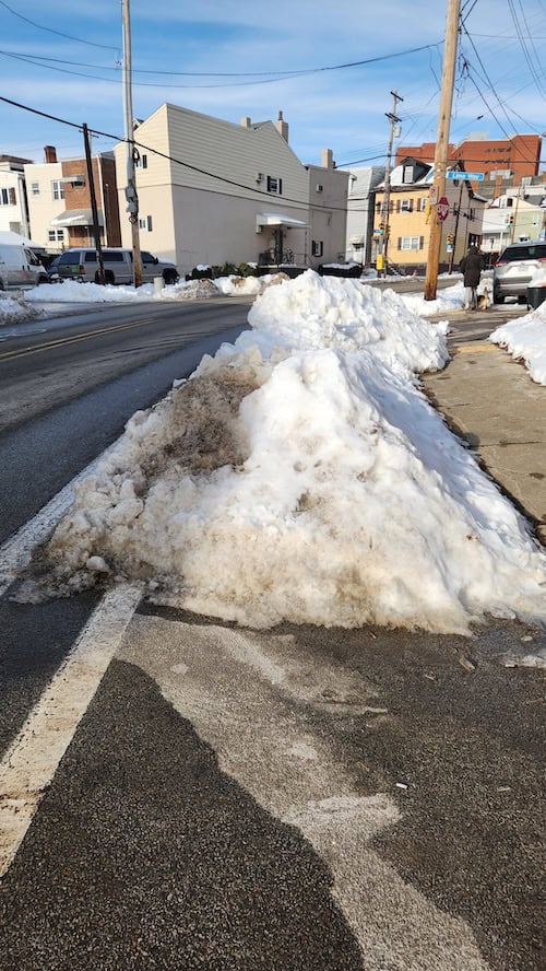 A giant mound of snow swallowing a bike lane, while the road where the cars drive is cleared and plowed. 