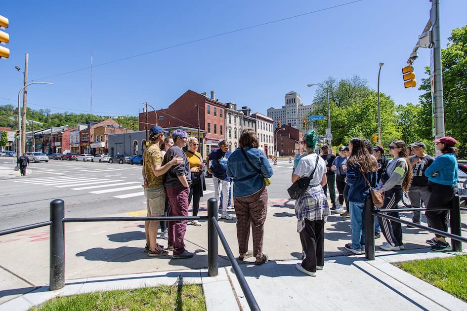 A group of about 25 people of all ages gather at a crosswalk in Pittsburgh's Northside on a sunny blue sky day 

