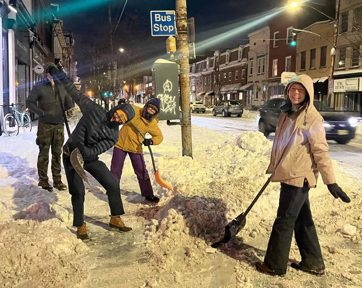 4 smiling friends in winter coats with snow shovels are shoveling a sidewalk on Liberty Ave at night time
