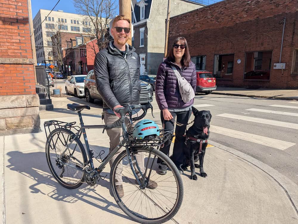 Seth and Amelie smiling together standing on the sidewalk with a bike and a black german shepherd guide dog. Seth is a a fair skinned man with red hair,  and Amelie is a fair skinned woman with medium length brown/auburn hair. They are both wearing sunglasses and looking happy on a sunny day. 