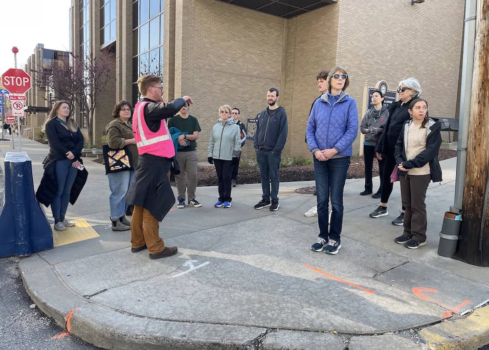 A group of 12 people gather at the corner of a sidewalk at Pitt. One person in a pink vest is explaining sidewalk safety while the others look onward.