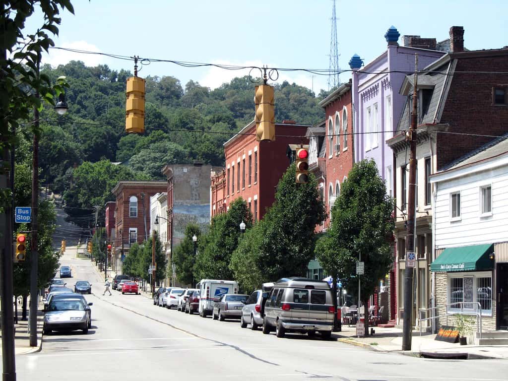 image of a street in West Pittsburgh with wide driving lanes, car parking on both sides of the street, and a bus stop