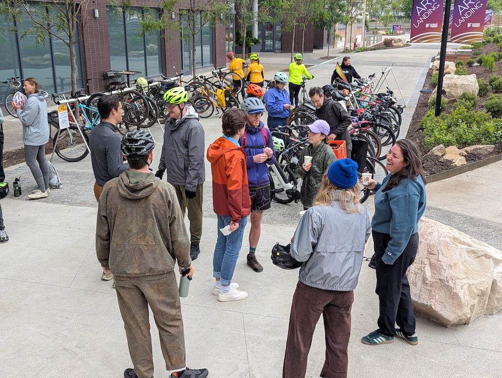 About 15 people gather in their colorful rain gear with lots of bikes and coffee cups, chatting and hanging out