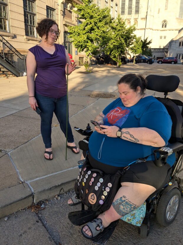 Person in wheelchair assessing a sidewalk with someone standing holding a tomato stake.