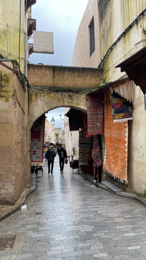 Two people walking under a stone archway past a colorful carpet market