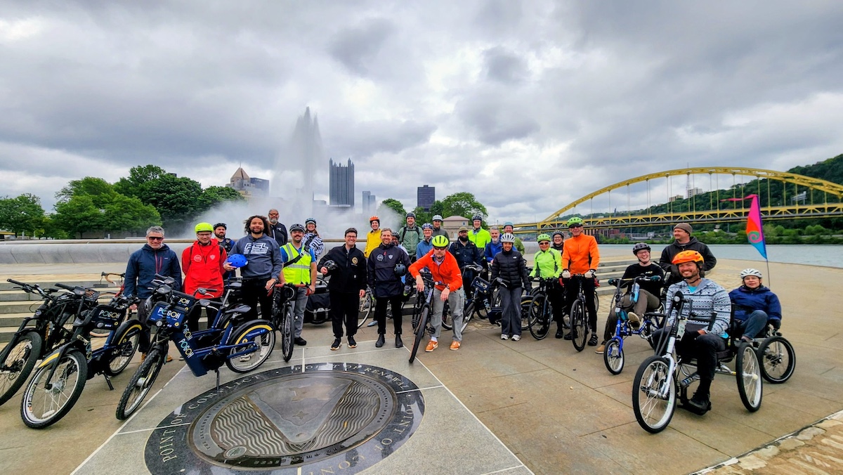 A group of 20 or 30 smiling bike rides,  with Mayor Corey O'Connor, stopping at The Point for a photo op, with city skyline and bridge in the background