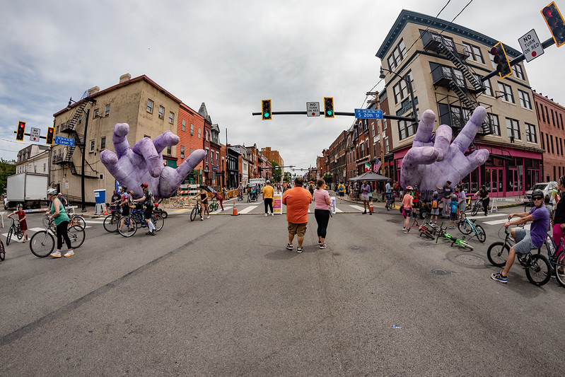 A photo of East Carson and 20th in Pittsburgh's South Side activated for OpenStreetsPGH, car-free, filled with 30 or so people, with giant purple hands art installation.