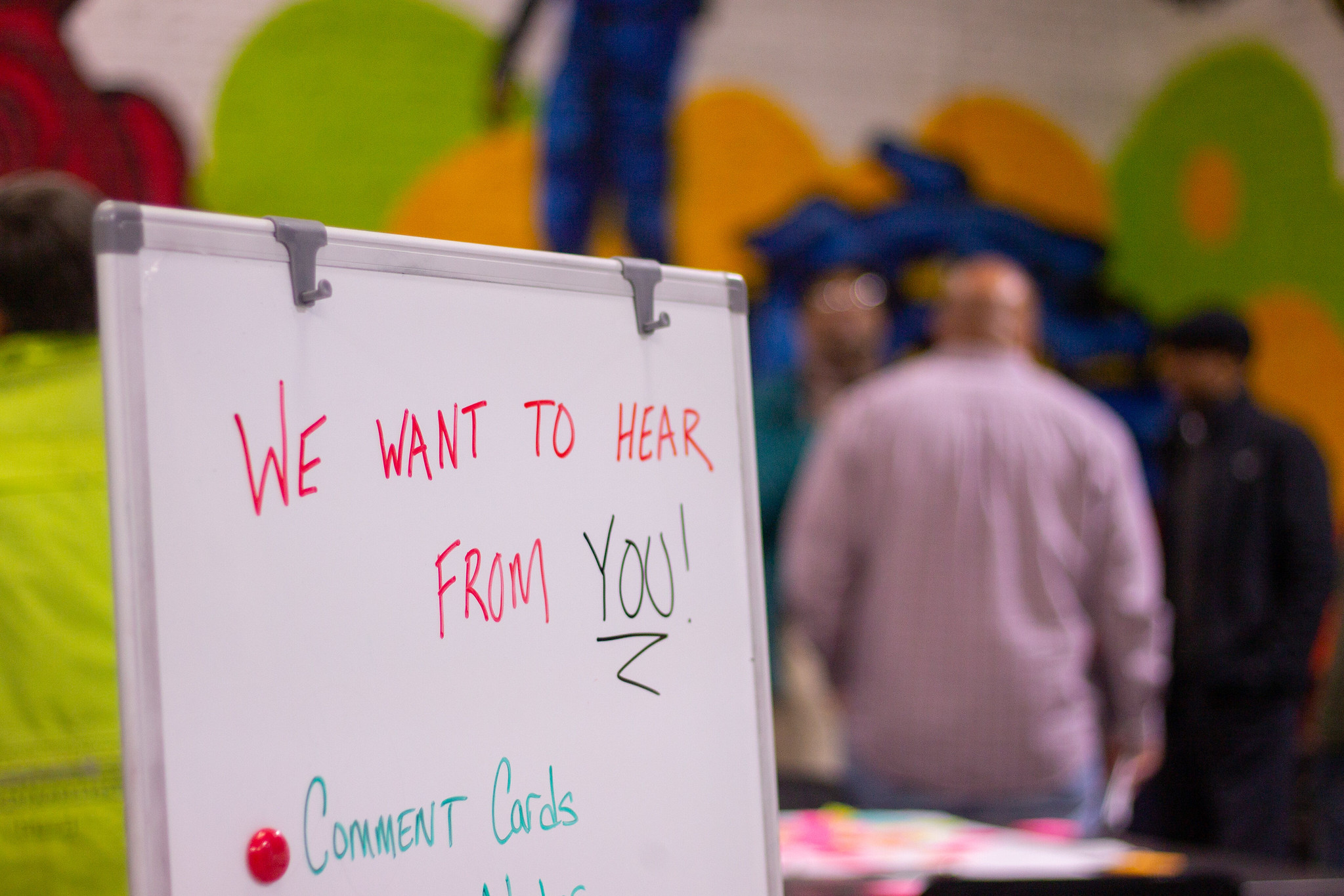 Picture of a whiteboard with the words "We want to hear from you" written in marker with people out of focus in the background.