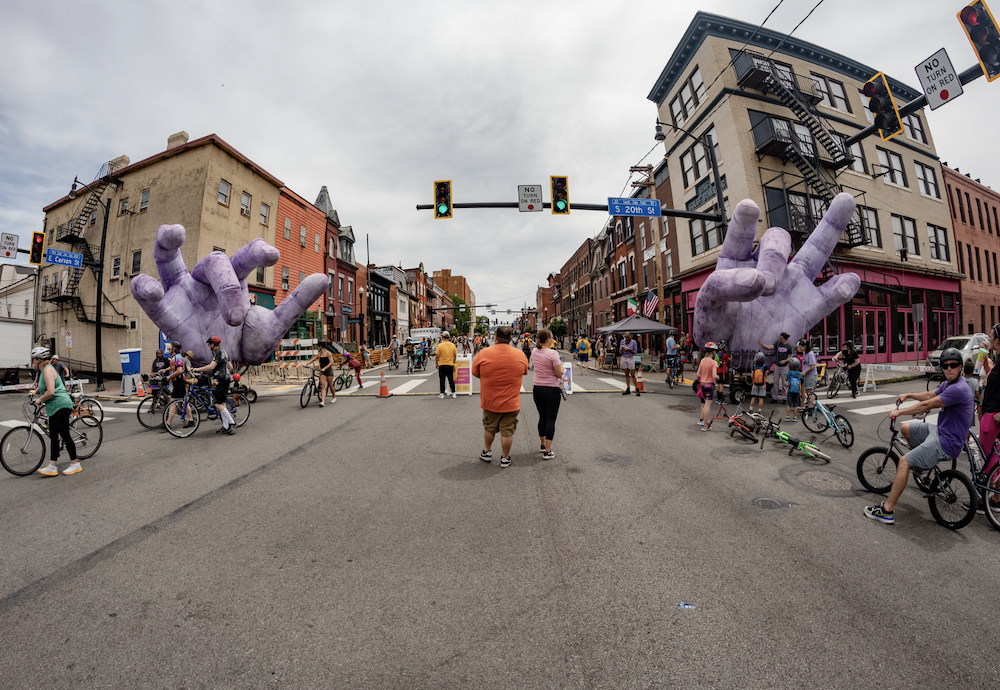 20 to 30 people of all ages walking and biking on E Carson St and S 20th S in Pittsburgh's South side on a sunny day