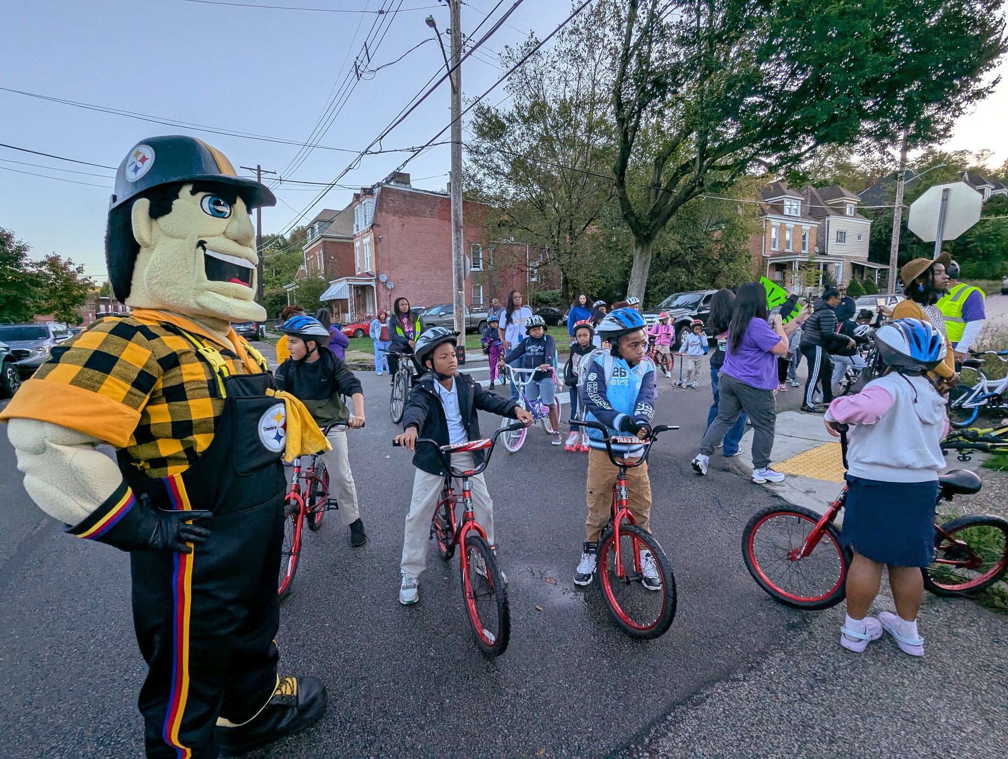 Steeler's Mascott, Steely McBeam, stands in the foreground. Behind him several dozen students sit astride bicycles with adult volunteers wearing neon vests. 