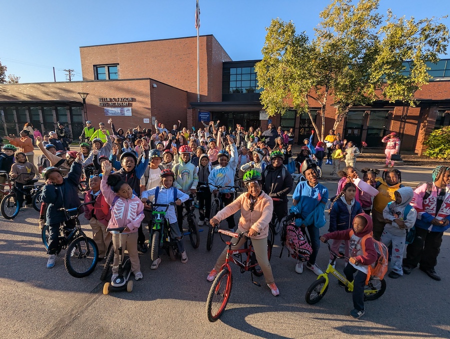 A group of over 50 students on bikes from ages 6 through 10 standing in front of Pittsburgh Faison school with their bikes and scooters, smiling and raising their hands above their heads in joy. 
