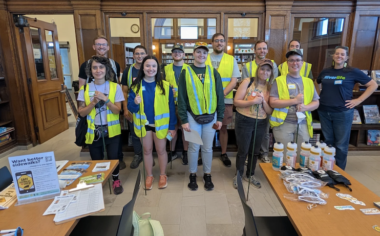 A group photo of 12 folks from the Safe Sidewalks Crew at the carnegiue library in Mount Washington. They are wearing hi-vis neon vests and holding tomato stakes, smiling and ready to record some sidewalks.