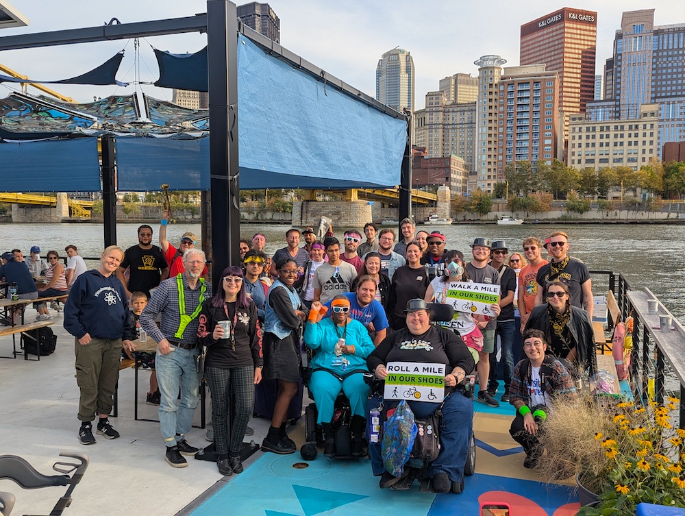 A big group photo at the Shore Thing with the city of Pittsburgh skyline behind the crowd. People in fun costumes holding signs that say "Roll a mile in our shoes", "walk a mile in our shoes."